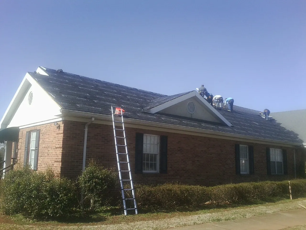 Crew tearing shingles off a brick ranch roof with a ladder set — Metal Roof Repair work by our crew in Mead