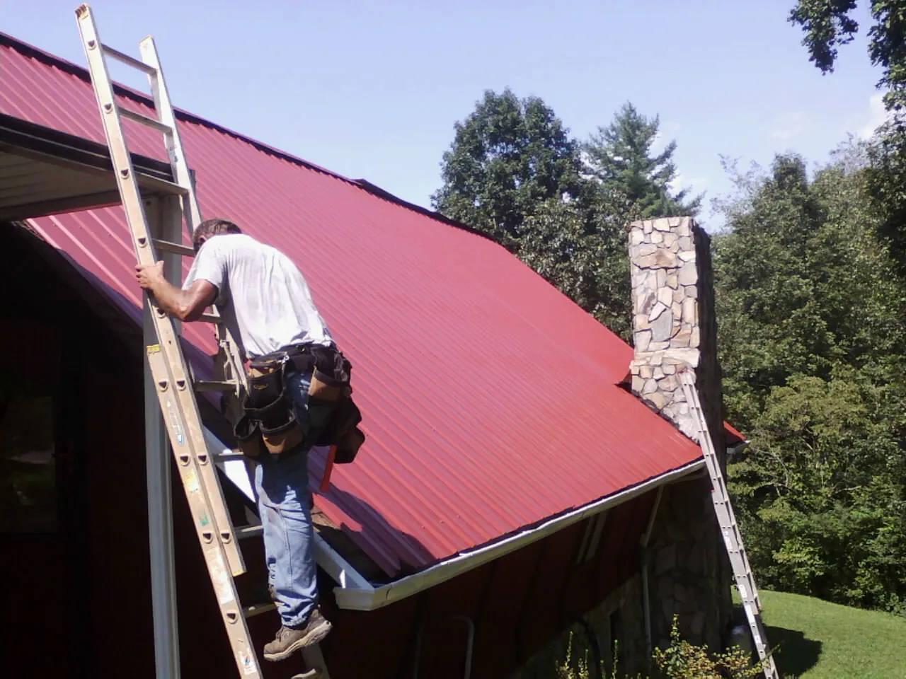 Roofer on a ladder beside a red metal roof and stone chimney — Soffit & Fascia Repair by Roofing Brothers in Mead